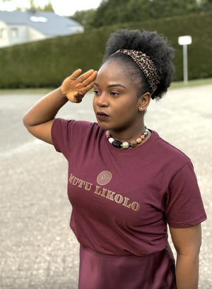 Femme portant un t-shirt bordeaux Mutu Likolo – Collection Résilience, effectuant un salut militaire, symbole de dignité et de force. | Woman wearing a burgundy Mutu Likolo T-shirt – Resilience Collection, giving a military salute, symbol of dignity and strength.